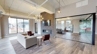 This contemporary single-floor commercial space is thoughtfully designed with a blend of modern aesthetics and functional versatility. The main lounge area serves as the centerpiece, featuring soaring ceilings with exposed structural elements, warm wood-look flooring, and a striking floor-to-ceiling stacked stone accent wall housing a linear fireplace and wall-mounted television. Statement pendant lighting, including a sputnik-style chandelier, adds a sophisticated ambiance throughout. Comfortable seating arrangements invite casual gatherings or relaxed collaboration.

Adjacent to the lounge, a glass-walled conference and dining area provides a bright, semi-private space outfitted with high-top tables and bar seating, ideal for meetings or group work sessions. Large windows along the exterior walls flood this area with abundant natural light and views of the surrounding neighborhood.

Full-height glass partition walls with sleek black metal framing define separate zones while maintaining an open, airy feel across the floor plan. A dedicated open flex space beyond the glass partitions offers additional room for various uses, with recessed lighting and the same warm wood-look flooring carried throughout.

A well-appointed kitchenette serves the space with dark wood cabinetry, light countertops, a stainless steel refrigerator, sink, and coffee station — ensuring convenience for daily use. A corridor connects to additional private rooms, completing this polished and highly functional commercial environment.