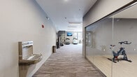 Located at 6325 Victoria Ave in St. Louis, Missouri, this well-appointed fitness center offers a comprehensive, single-floor workout facility designed to support a full range of exercise routines. The space features wood-look flooring throughout, neutral gray walls, and bright flush-mounted lighting that creates a clean, energizing atmosphere.

The cardio zone is equipped with multiple treadmills, an elliptical, a stationary bike, and a rowing machine, all positioned near large windows that allow natural light to fill the space and provide views of the surrounding area. A wall-mounted flat-screen television is conveniently placed for entertainment during workouts.

The strength training area includes a cable and pulley multi-functional machine, a power rack with an adjustable bench, a dedicated dumbbell rack with a full range of free weights, and a step platform for functional training. Floor-to-ceiling mirrors line the strength section, enhancing both the sense of space and utility for form-checking during exercise.

A wide corridor connects the main workout floor to a glass-enclosed studio space, offering flexibility for group fitness or private training sessions. A water fountain is accessible near the entrance for added convenience. The overall layout is open and spacious, allowing for comfortable movement between equipment zones while maintaining a modern, professional aesthetic.