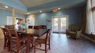 This single-floor community amenity space is thoughtfully laid out to serve a variety of recreational and social needs. The fitness center is outfitted with multiple treadmills and cardio machines, accented by bold orange and blue walls, recessed and overhead lighting, and windows that allow natural light to filter in. The lounge area is a warm and inviting retreat, anchored by a sleek fireplace framed by floor-to-ceiling windows dressed with sheer white curtains. The rich earth-toned walls and generous glazing offer expansive outdoor views of surrounding greenery, creating a bright and tranquil atmosphere. The social dining area features tile flooring, soft blue walls, recessed lighting, and a large wooden dining table with seating for multiple guests. French doors open to the exterior, enhancing the sense of openness. The full kitchen is equipped with stainless steel appliances, including a range, microwave, and refrigerator, complemented by light wood cabinetry, dark granite countertops, and a decorative tile backsplash. A peninsula counter adds additional workspace. Together, these spaces form a cohesive, functional, and welcoming environment suited for gatherings, fitness, and everyday community life.