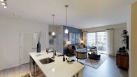 This 2-bedroom, 2-bathroom residence is thoughtfully designed with an open-concept layout that seamlessly connects the kitchen and living areas. The kitchen features a large island with a bright white countertop, an undermount sink with a modern gooseneck faucet, and dark cabinetry beneath. Pendant lights hang elegantly above the island, providing focused task lighting while complementing the overall aesthetic.

The living area flows naturally from the kitchen, anchored by a neutral-toned area rug and furnished with a mix of seating arrangements centered around a round ottoman-style coffee table. A bold slate-blue accent wall adds depth and visual interest to the space, serving as a backdrop for framed artwork and a console table with ambient lighting.

Floor-to-ceiling sliding glass doors flood the living area with natural light and open to a balcony with urban views beyond. White bifold doors, visible near the kitchen, likely lead to a laundry or storage closet. The overall ambiance is modern and sophisticated, with a cohesive neutral palette punctuated by rich tones and contemporary furnishings throughout.