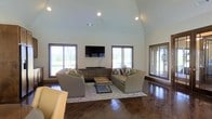 This single-story space is defined by its striking vaulted ceiling, which rises dramatically to create an airy, open atmosphere in the main living area. The ceiling's sharp pitch is complemented by recessed lighting and a ceiling fan, ensuring the space is well-lit and comfortable throughout the day.

The living area is anchored by rich, dark hardwood flooring that extends throughout the room, providing a warm contrast to the bright white walls and high ceiling. A wall-mounted flat-screen television serves as a focal point, positioned above a low media console between two generously sized windows. The seating arrangement includes two neutral-toned sofas and an armchair, accented with green and patterned throw pillows, all centered around a rectangular wooden coffee table atop a decorative area rug.

Multiple windows fitted with horizontal blinds line the walls, inviting abundant natural light while offering views of the surrounding greenery. The overall ambiance is clean, modern, and welcoming, with a careful balance of functional furnishings and architectural character that makes the space feel both spacious and comfortable.