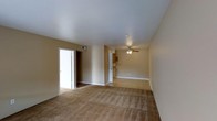 This single-floor residential space presents a straightforward and functional layout designed for comfortable everyday living. The main living area is generously sized, finished with wall-to-wall plush carpeting in a warm neutral tone that extends throughout the space, adding softness underfoot and contributing to an inviting atmosphere. Smooth, white ceilings are fitted with a ceiling fan, providing both ambient lighting and airflow to keep the space comfortable year-round. The walls are finished in a muted beige tone, offering a neutral backdrop that suits a variety of interior styles. An open passageway connects the living area to what appears to be a kitchen, where cabinetry in a warm wood finish is visible, hinting at a practical cooking space. A doorway off the main living area leads to additional rooms, suggesting a well-organized floor plan that separates living and private spaces effectively. Recessed ventilation is incorporated into the upper wall, supporting efficient climate control throughout. The overall ambiance is clean, understated, and move-in ready, making this a practical residential space with a calm, neutral aesthetic well-suited for personalization.