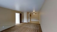 This single-floor residential space presents a straightforward and functional layout designed for comfortable everyday living. The main living area is generously sized, finished with wall-to-wall plush carpeting in a warm neutral tone that extends throughout the space, adding softness underfoot and contributing to an inviting atmosphere. Smooth, white ceilings are fitted with a ceiling fan, providing both ambient lighting and airflow to keep the space comfortable year-round. The walls are finished in a muted beige tone, offering a neutral backdrop that suits a variety of interior styles. An open passageway connects the living area to what appears to be a kitchen, where cabinetry in a warm wood finish is visible, hinting at a practical cooking space. A doorway off the main living area leads to additional rooms, suggesting a well-organized floor plan that separates living and private spaces effectively. Recessed ventilation is incorporated into the upper wall, supporting efficient climate control throughout. The overall ambiance is clean, understated, and move-in ready, making this a practical residential space with a calm, neutral aesthetic well-suited for personalization.
