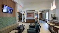 This single-floor lobby presents a refined and contemporary design, combining functional and aesthetic elements seamlessly. The space is anchored by a prominent wooden reception desk, accented with a stone or tile facade, and topped by a bold abstract painting in warm earth tones of orange, red, and brown — serving as a striking focal point for the room.

The ceiling is elevated with clean crown molding details, contributing to an open and airy atmosphere. Recessed and wall-mounted lighting supplements the natural light filtering in through large glass doors at the rear, offering views of an outdoor area beyond.

The seating area in the foreground features dark wood-framed armchairs with teal upholstery arranged around a low coffee table adorned with decorative objects, conveying a polished, hospitality-oriented atmosphere. The flooring transitions between light tile and patterned area rugs, adding visual texture throughout.

Additional functional spaces are accessible through corridors on either side, with glimpses of a lounge or common area and what appears to be a dining space visible to the right. Decorative sculptures and accent pieces throughout enhance the space's curated, upscale character.