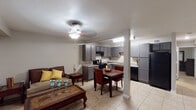 Located at 6425 S Gessner Rd in Houston, TX 77036, this single-floor unit offers a functional and well-organized layout suited for comfortable everyday living. The open-concept main area seamlessly integrates the living room, dining space, and kitchen, creating a connected and airy feel. The living area features light tile flooring, a ceiling fan with light fixture, and warm neutral walls, while yellow accent curtains add a touch of color. A dining table with seating sits centrally, bridging the living and kitchen zones naturally. The kitchen is well-appointed with gray shaker-style cabinetry extending to the ceiling, speckled laminate countertops, a double-basin stainless steel sink, black appliances including an electric range and refrigerator, and a built-in dishwasher — providing ample storage and prep space. The bedroom continues the tile flooring theme and includes a ceiling fan, a closet, and an attached hallway leading to the bathroom. The bathroom is equipped with accessibility features including grab bars near the toilet and shower area, a shower with curtain enclosure, and a vanity with countertop. Throughout the unit, the consistent tile flooring, neutral color palette, and practical fixtures create a cohesive and low-maintenance living environment.
