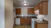 This single-floor space centers around a practical and well-appointed kitchen that serves as its standout feature. The kitchen is designed in an L-shaped layout, maximizing efficiency and workflow. Warm honey-toned wood cabinetry lines the walls both above and below the countertops, providing generous storage throughout. The dark laminate countertops offer a grounding contrast to the lighter cabinetry, creating a cohesive and classic look. A double-basin sink is positioned centrally along the lower cabinetry, complemented by a built-in range hood above the cooking area. The kitchen is fully equipped with white appliances, including a coil-burner electric range, a dishwasher, and a full-sized refrigerator, making it ready for everyday use. Durable tile flooring in a earthy, mottled pattern runs throughout, offering both practicality and visual texture underfoot. Overhead, a flush-mount ceiling light provides consistent illumination across the space. The overall ambiance is functional and comfortable, with a straightforward, no-frills design that prioritizes usability and practicality for everyday living.
