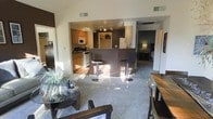 This single-floor space is designed with an open-concept layout that seamlessly connects the living, dining, and kitchen areas, creating a cohesive and functional environment. The kitchen features warm honey-toned cabinetry, a gas range, and a built-in microwave, complemented by a dark-finished breakfast bar with stylish bar stools — an ideal spot for casual dining or entertaining. Track lighting illuminates the kitchen area, while vaulted ceilings add a sense of vertical spaciousness to the open living zone. The living area is finished with plush carpeting and benefits from generous natural light streaming through the space, creating a bright and welcoming atmosphere. A glass-topped coffee table and seating area anchor the living space, while a dining table with high-back chairs sits conveniently nearby. A hallway visible from the main area leads to additional rooms, including what appears to be a bedroom with a lamp-lit nightstand. The contrast between the tiled kitchen flooring and the carpeted living area subtly defines each zone while maintaining the open flow throughout.