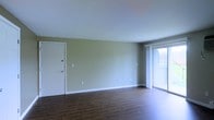 This single-floor space offers a clean, functional layout centered around an open living area. The room is finished with rich, dark hardwood-style flooring that contrasts neatly with the soft, neutral green-toned walls and crisp white baseboards and trim throughout. A large sliding glass door dominates one wall, allowing an abundance of natural light to pour into the space and providing a seamless visual connection to the outdoors. The interior entry door is finished in white, consistent with the clean, minimal aesthetic carried throughout the space. The walls feature standard outlets and light switch placements, reflecting a practical, move-in-ready design. The ceiling is smooth and unadorned, reinforcing the simple, understated character of the space. Overall, the space presents a bright, open atmosphere with a neutral palette that offers flexibility for a variety of furnishing and decorating styles. The combination of durable flooring, ample natural light from the sliding glass door, and well-maintained finishes makes this a versatile and welcoming environment.