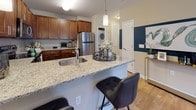 This single-floor residence offers a thoughtfully designed layout that balances functionality and style throughout its living spaces. The kitchen serves as a central highlight, showcasing rich warm-toned cabinetry, speckled granite countertops, and a full suite of stainless steel appliances including a range, microwave, refrigerator, and dishwasher. A peninsula with an integrated sink and pendant lighting overhead provides additional prep space and casual bar seating, making it ideal for both cooking and entertaining. Conveniently tucked nearby is an in-unit stacked washer and dryer. Light hardwood-style flooring runs throughout the main living areas, adding warmth and cohesion to the space. The living room is a comfortable retreat, featuring neutral walls and ample room for a sectional sofa arrangement, with decorative drapery adding visual interest. The bedroom is well-proportioned, bathed in natural light through dual windows dressed with breezy curtains, and accented by a bold feature wall. The bathroom is clean and functional, finished with white tile flooring, a tub-shower combination, and a warm wood vanity with a light countertop. Overall, the space exudes a modern yet welcoming ambiance.