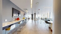 Located at 1457 North Halsted Street in Chicago's vibrant Lincoln Park/River North corridor, this sophisticated amenity lounge occupies a single, generously proportioned floor with an open, flowing layout. The space is defined by sweeping floor-to-ceiling curtain-wall windows along multiple elevations, flooding the interior with abundant natural light and offering panoramic views of the Chicago skyline and surrounding cityscape.

Large-format stone-effect tile flooring runs continuously throughout, lending a clean, upscale aesthetic that unifies the various functional zones. Structural columns are finished in smooth concrete and grey tile cladding, serving as subtle architectural anchors within the open plan.

The space is organized into several distinct yet connected areas. A long bar-height counter with modern stools along one wall accommodates casual dining and socializing, complemented by a wall-mounted display screen. Round café tables with contemporary chairs provide additional flexible seating throughout the central area. Intimate lounge vignettes feature plush upholstered armchairs and sofas arranged on area rugs, ideal for relaxed gatherings. A curved, glass-enclosed alcove offers a serene seating nook with unobstructed views of an adjacent outdoor terrace and the city beyond.

Recessed linear lighting in the white ceiling adds a polished finishing touch. Potted ornamental grasses and plants are distributed throughout, introducing natural warmth to the modern, minimalist palette.