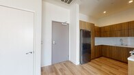 This contemporary single-floor unit offers a thoughtfully designed layout with clean, modern finishes throughout. The entryway opens to light wood-toned flooring that extends seamlessly through the main living areas, creating a warm and cohesive aesthetic against the crisp white walls.

The kitchen is a standout feature, outfitted with rich wood-grain cabinetry that extends to the ceiling, providing ample storage. White countertops and a tile backsplash offer a clean contrast, while stainless steel appliances — including a full-size refrigerator and microwave — complete the functional and stylish space.

The living area is bright and open, with elevated ceilings that enhance the sense of space. Floor-to-ceiling glass doors lead to a private balcony, offering panoramic city views that bring in abundant natural light. A dedicated laundry closet with a stacked washer and dryer unit adds everyday convenience.

The bedroom features plush carpet flooring and a large window framing expansive city views, creating a calm, light-filled retreat. The full bathroom is appointed with warm wood vanity cabinetry, large-format tile flooring, and a tub and shower combination with elegant marble-style tile surround, lending a refined, spa-like quality to the space.