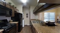 This single-story space centers around a well-appointed galley-style kitchen that maximizes efficiency through its narrow, linear layout. The kitchen is finished with granite countertops that provide ample prep space along both sides, complemented by rich, dark wood cabinetry that extends from lower storage units to upper cabinets mounted near the ceiling, offering substantial storage capacity. Black appliances, including a refrigerator and range, coordinate with the darker cabinetry tones for a uniform aesthetic.

A double-basin stainless steel sink is positioned along the counter, served by a standard faucet fixture. Upper cabinets with glass panel inserts add a subtle decorative detail to the otherwise functional space. Dark hardwood flooring runs throughout, adding warmth and visual continuity.

At the end of the kitchen corridor, a dedicated laundry area accommodates a stacked washer and dryer unit, a practical in-unit convenience. Neutral beige walls throughout keep the interior feeling open despite the compact layout. Natural light filters in from an adjacent room visible at the far end, suggesting a connected living or dining area nearby.