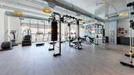 This contemporary fitness facility is thoughtfully designed to accommodate a wide range of workout styles and training goals. The space is organized into distinct functional zones, each outfitted with professional-grade equipment. The main workout floor features an impressive array of cardio machines, including treadmills and stationary bikes, alongside strength training stations with cable systems, benches, and a full dumbbell rack. A dedicated functional training area includes kettlebells of varying weights neatly arranged on a tiered rack, medicine balls, and a heavy punching bag suspended from a freestanding rig. A separate, glass-enclosed studio provides a quieter space for focused training or classes, offering visual connectivity to the main floor while maintaining acoustic separation. The interior design strikes a balance between industrial and modern aesthetics, with exposed ductwork running along the high ceilings, warm wood-look flooring, and crisp white walls. A large, vibrant sports-themed mural energizes the main workout area, while strategically placed mirrors expand the sense of space. Wall-mounted flat-screen displays and pendant lighting with gold accents add polished finishing touches throughout, creating an inviting and motivating environment for all fitness levels.