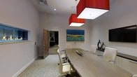 Located at 201 Willett Ave in Port Chester, New York, this single-floor commercial space offers a polished and sophisticated environment designed for professional use. The centerpiece of the space is a sleek conference room that seamlessly blends modern design with functional elegance. A striking built-in linear fireplace spans a large portion of the wall, providing a dramatic focal point that adds warmth and visual interest to the room. The conference area is anchored by a long, wood-grain table surrounded by white ergonomic chairs, creating a refined setting ideal for meetings and collaborative work. Overhead, bold red square pendant light fixtures serve as a defining design element, lending a vibrant contrast to the neutral, light-toned walls and carpeted floors. Recessed lighting throughout ensures the space is well-illuminated, while a wall-mounted display screen adds modern utility. Warm wood-finished doors complement the contemporary aesthetic, and the open sightlines into adjacent workspace areas suggest a thoughtfully connected floor plan. The overall ambiance is one of understated luxury — professional yet inviting, with carefully curated design details that elevate the standard office environment.