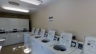 This single-floor shared laundry facility is designed for functional, efficient use. A row of top-loading washing machines lines the center of the room, each equipped with individual digital payment and control panels, making operation straightforward for users. Along the back wall, stacked washer-dryer units provide additional laundry capacity, maximizing the use of available space. The room features neutral beige walls and is illuminated by overhead fluorescent lighting, creating a clean, well-lit environment. The flooring is light-toned and easy to maintain, contributing to the overall tidy appearance of the space. A posted notice on the wall provides users with guidelines and instructions for proper use of the machines. The layout is open and accessible, allowing multiple users to navigate the space comfortably at the same time. The facility appears well-maintained, with all equipment in clean condition, reflecting a practical and organized shared amenity suited for residential or commercial use.