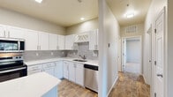 This single-floor residence offers a thoughtfully designed layout with contemporary finishes throughout. The kitchen is a standout feature, showcasing crisp white shaker cabinetry with brushed nickel hardware, light grey subway tile backsplash, and bright white countertops that extend to a generously sized center island. Stainless steel appliances — including a French door refrigerator, electric range, built-in microwave, and dishwasher — complete the space, while wood-look flooring adds warmth underfoot. A decorative wine rack built into the upper cabinetry adds a charming touch. The living area is open and airy, featuring the same durable wood-look flooring, a corner fireplace as a focal point, and a window that invites natural light and outdoor views. The bedrooms are carpeted for comfort, with multiple windows offering tree-lined views and ample wall space for furnishing flexibility. The bathroom is clean and functional, featuring a white vanity with an undermount sink, a large mirror, and a walk-in shower. A spacious walk-in closet with wire shelving provides excellent storage. Neutral wall tones and consistent finishes throughout give the space a cohesive, modern aesthetic.