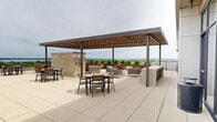 This rooftop terrace is a thoughtfully designed outdoor amenity space offering a rich variety of settings for relaxation, dining, and socializing. The terrace is defined by its expansive open-air layout, finished with large-format pavers that provide a clean, contemporary foundation throughout.

A standout feature is the covered pergola area, constructed with warm wood slats and steel supports and strung with ambient café lighting. Beneath it, plush lounge seating is arranged around a stone fireplace column, creating an inviting gathering spot. Surrounding raised wooden planter beds filled with ornamental grasses and greenery add a natural, garden-like quality to this zone.

Adjacent to the pergola, an open sun deck hosts multiple chaise lounges positioned against a dramatic floor-to-ceiling glass curtain wall, which separates the outdoor terrace from an interior amenity space and maximizes transparency between the two. Additional open-air dining areas feature durable table-and-chair sets arranged to take full advantage of the panoramic views.

The terrace's perimeter is lined with sleek glass railings, offering unobstructed sightlines across a lush tree canopy, the surrounding urban neighborhood, and a broad lake stretching to the horizon. The elevated vantage point provides sweeping 360-degree views, making this rooftop terrace a premier outdoor destination within the building.