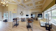 Located in Pinellas Park, Florida, this single-story community clubhouse is designed to serve as a central gathering hub for residents. The main lounge greets visitors with soaring coffered ceilings, warm yellow walls, and an elegant chandelier that casts a welcoming glow over light hardwood floors. Generously sized windows and French doors flood the space with natural light while offering direct views of the outdoor pool area, palm trees, and landscaped grounds beyond. High-top tables and bar-style seating are arranged throughout the open lounge, creating a relaxed yet social atmosphere. A dedicated café lounge area features a bold teal pendant fixture, a built-in coffee station with dark cabinetry, a wall-mounted television, and casual dining tables—ideal for a morning coffee or informal gathering. The clubhouse also includes a full kitchen accessible through an arched entryway, appointed with dark cabinetry, granite countertops, stainless steel appliances, and a large kitchen island with bar seating. Throughout the space, tasteful wall décor, sculptural accents, and consistent hardwood-style flooring tie the areas together cohesively. The overall ambiance is bright, modern, and inviting, reflecting a thoughtfully designed space built for community living.