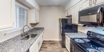 Located at 2100 Wilson Rd in Knoxville, Tennessee, this single-floor unit offers a well-organized layout with a clean, modern aesthetic throughout. The galley-style kitchen is efficiently designed with white cabinetry, granite-style countertops, a stainless steel sink, and black appliances including an electric range, refrigerator, and over-the-range microwave. A window with blinds allows natural light to brighten the space. Dark hardwood-style flooring runs through the kitchen and main living areas, providing a warm and cohesive look. The living room is a generously proportioned space featuring a fireplace with a classic white mantle, making it a natural focal point of the room. A hallway connects the main living areas to the additional rooms, one of which features plush carpeting and neutral wall tones. The bathroom is well-appointed with a full tub and shower combination, a granite-topped vanity with updated cabinetry, and hardwood-style flooring that continues the design language of the rest of the unit. Textured ceilings and clean white trim are consistent throughout, contributing to a tidy and cohesive interior. The overall ambiance is bright, neutral, and move-in ready.