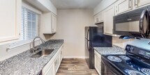 Located at 2100 Wilson Rd in Knoxville, Tennessee, this single-floor unit offers a well-organized layout with a clean, modern aesthetic throughout. The galley-style kitchen is efficiently designed with white cabinetry, granite-style countertops, a stainless steel sink, and black appliances including an electric range, refrigerator, and over-the-range microwave. A window with blinds allows natural light to brighten the space. Dark hardwood-style flooring runs through the kitchen and main living areas, providing a warm and cohesive look. The living room is a generously proportioned space featuring a fireplace with a classic white mantle, making it a natural focal point of the room. A hallway connects the main living areas to the additional rooms, one of which features plush carpeting and neutral wall tones. The bathroom is well-appointed with a full tub and shower combination, a granite-topped vanity with updated cabinetry, and hardwood-style flooring that continues the design language of the rest of the unit. Textured ceilings and clean white trim are consistent throughout, contributing to a tidy and cohesive interior. The overall ambiance is bright, neutral, and move-in ready.
