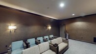 This elegantly appointed private screening room delivers an immersive cinematic experience within a refined, upscale setting. The space is dressed in deep, richly textured dark walls that absorb light and enhance the theater atmosphere, complemented by crown molding and soft, patterned carpet flooring throughout.

Seating is arranged in a tiered layout, combining plush light-toned sofas and individual lounge chairs in a soft blue-gray upholstery, all framed by dark leather armrests and equipped with built-in cupholders — thoughtfully designed for comfort during extended viewing. The focal point of the room is a large projection screen framed by floor-to-ceiling two-tone drapes in neutral tones, flanked by wall sconces that cast a warm, ambient glow.

Recessed ceiling lighting and additional wall-mounted sconces provide layered illumination that can be adjusted to suit the viewing environment. A discreet wet bar area features a countertop sink, cabinetry with drawers, and an under-counter mini refrigerator, making the space well-suited for entertaining. A stainless steel waste bin and a keypad-entry door complete the thoughtful, self-contained design of the room, ensuring a seamless and private screening experience.