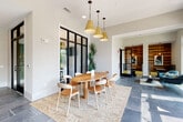 This thoughtfully designed single-story space combines contemporary aesthetics with functional versatility. Large-format slate tile flooring flows throughout, unifying distinct zones while lending an air of sophistication. Black-framed floor-to-ceiling windows and glass-panel doors are defining architectural elements, flooding the interior with natural light and framing lush outdoor greenery.

A central dining area features a rounded wood table with modern chairs, anchored by a vintage-style area rug and illuminated by a cluster of warm gold pendant lights. Adjacent to this, a comfortable lounge area offers teal upholstered chairs positioned beside tall picture windows that overlook a landscaped exterior, ideal for informal gatherings or relaxation.

A dedicated co-working zone provides a long counter-height work surface lined with cushioned stools, accented by brass swing-arm wall sconces and curated blue-toned artwork. Private enclosed offices, accessible through glass-panel sliding doors, offer quiet focused workspaces. A warm wood-clad feature wall adds textural contrast near the entryway. Throughout, the space balances openness with privacy, maintaining a bright, modern, and welcoming ambiance.