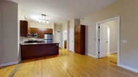 This single-floor residence offers a cohesive and functional layout unified by warm hardwood flooring that extends throughout every room. The open-concept kitchen serves as a central hub of the space, featuring rich medium-toned wood cabinetry, granite countertops, a large island with a double undermount sink, and stainless steel appliances including a refrigerator, range, and built-in microwave. Pendant lighting above the island adds a refined finishing touch. The kitchen flows seamlessly into the open living area, creating an expansive, connected feel. Multiple bedrooms provide versatile living options; one features a pair of large windows that invite natural light and offer views of the surrounding urban architecture, while another includes a tall built-in wood cabinet for added storage. A smaller room with a single window offers a cozy, flexible space ideal for a bedroom or home office. The bathroom is well-appointed with warm wood vanity cabinetry, granite countertops, an undermount sink, and classic black hardware accents. Throughout the residence, neutral wall tones in soft beige and warm greige complement the natural wood floors, creating a consistent and inviting ambiance. Paneled white doors with black hardware add a clean, classic character to the overall design.
