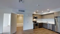 This single-floor space showcases a clean, contemporary design characterized by an open layout that connects the main living area to the kitchen. The kitchen is equipped with stainless steel appliances, including an electric range, over-the-range microwave, and a sink, all framed by a two-tone cabinetry scheme featuring upper light wood-tone cabinets and darker lower cabinets. A white tile backsplash adds a crisp, clean contrast to the cabinetry. Recessed lighting illuminates the kitchen area, while the broader living space benefits from ceiling-mounted HVAC vents, ensuring climate control throughout. Wood-look plank flooring extends continuously across the space, adding warmth and cohesion to the interior. Directly accessible from the main area is a bathroom, which features a combination shower and bathtub with tile surround, a toilet, and overhead lighting. The white walls and smooth ceilings throughout the unit reinforce a minimalist aesthetic, creating a bright and functional environment well-suited for everyday living.