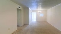 This single-floor residence offers a clean, neutral aesthetic with a welcoming open layout. The main living area is finished with plush wall-to-wall carpeting in a warm beige tone, creating a comfortable and cohesive feel underfoot. Textured popcorn ceilings add a classic touch overhead, complemented by crisp white crown molding that frames each room with a refined architectural detail. A standout feature of the dining or living area is an elegant chandelier that serves as a focal point, lending a touch of sophistication to the space. Natural light flows generously through bright doorways and windows, enhancing the airy ambiance throughout. The open floor plan allows for a seamless transition between the living and dining areas, offering flexibility in how the space can be arranged and used. A framed wall nook or pass-through adds a subtle architectural accent, while multiple doorways lead to additional rooms and closet storage, suggesting practical and functional utility. The overall palette of soft whites and beiges creates a calm, move-in-ready atmosphere that is both versatile and inviting.