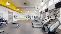 Located at 825 Cleveland Ave in Columbus, Ohio, this well-appointed fitness center offers a spacious, single-floor layout designed to support a full range of workout routines. The space features soaring ceilings with exposed ductwork and pendant lighting, creating an open, industrial-modern aesthetic. Bold yellow accent walls contrast with crisp white surfaces, giving the facility an energetic and motivating atmosphere.

Natural light pours in through a generous row of clerestory and lower windows along multiple walls, keeping the space bright and airy throughout the day. The durable speckled rubber flooring provides both safety and comfort underfoot across the entire workout area.

The cardio zone is well-equipped with multiple treadmills, elliptical trainers, and a stair climber, each positioned in front of wall-mounted flat-screen televisions for an engaging workout experience. The strength training area features a comprehensive dumbbell rack, adjustable weight benches, a cable resistance machine, and additional free weight equipment. A dedicated stretching and functional training zone includes stability balls and yoga accessories, complemented by a large wall mirror.

A utility area with yellow cabinetry and a sink offers added convenience. Eye-catching wall art adds a creative, urban touch to the overall design, making this fitness center both functional and visually dynamic.