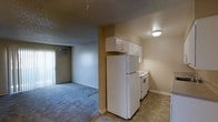 This single-floor unit offers a straightforward and functional layout designed for comfortable everyday living. The living area is finished with wall-to-wall carpet and features a wall-mounted heating unit, providing a warm and practical space. A window-mounted air conditioning unit ensures year-round climate control. The textured popcorn ceiling is consistent throughout, lending a classic residential feel to the interior.

The kitchen is designed in a galley configuration, maximizing efficiency within a compact footprint. White cabinetry lines both sides of the kitchen, offering generous storage space above and below the countertops. The counters feature a speckled laminate finish, complementing the white appliances, which include a refrigerator and an electric range with oven. A stainless steel sink is positioned along one wall, and tile flooring runs throughout the kitchen area, providing a durable and easy-to-clean surface.

An open transition connects the kitchen to the main living space, where the carpet begins. A hallway, illuminated by a wall-mounted light fixture, leads to additional rooms beyond, offering privacy and separation between living and private areas. Overall, the space presents a clean, modest, and move-in ready interior suited for practical, day-to-day use.