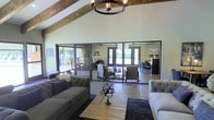 This single-floor space showcases a thoughtfully curated rustic-modern aesthetic that seamlessly blends warmth and sophistication. The main living area immediately draws the eye upward to its dramatic vaulted ceiling, where rich, exposed wooden beams run the length of the roofline, adding architectural character and a sense of organic texture. A wrought-iron chandelier with candle-style lighting anchors the space from above, complementing the rustic elements throughout.

The living area is furnished with tufted gray sofas arranged around a reclaimed wood coffee table, accented by a decorative armillary sphere centerpiece that adds a refined, classic touch. Underfoot, herringbone-patterned hardwood flooring lends an elegant structure to the open layout.

A defining feature of this space is the floor-to-ceiling steel-framed glass partition wall, which divides the main living area from adjacent rooms while maintaining an open, airy feel and allowing natural light to flow freely throughout. Beyond the glass, a dining area and additional seating areas are visible, each benefiting from abundant natural light streaming in through large glass doors and windows that open to the outdoors. The overall ambiance is bright, welcoming, and cohesive, balancing rustic materials with clean, modern lines.