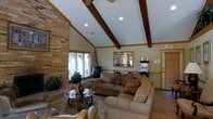 This inviting single-floor space makes an immediate impression with its dramatic vaulted ceilings adorned with rich, dark-stained exposed wood beams that draw the eye upward and lend a sense of rustic elegance. Recessed lighting and a ceiling fan complement the natural light that streams in through large windows, keeping the interior bright and airy. A floor-to-ceiling brick accent wall anchors the living area, adding texture, warmth, and a classic architectural character that serves as a defining focal point of the room. The living area is generously proportioned, offering ample space for comfortable seating arrangements. Plush, neutral-toned sofas and accent pillows create a cozy gathering space centered around a glass-top coffee table with a decorative iron base. A decorative mirror adorns the wall, reflecting light and adding depth to the room. Warm-toned walls and wood accents throughout contribute to the cohesive, welcoming ambiance. The space flows naturally, with clean sightlines that emphasize its openness. Overall, the design blends traditional charm with casual comfort, making it a functional and aesthetically appealing environment suited for both relaxed everyday living and entertaining.