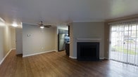 This single-floor residence offers a well-designed open layout that combines comfort and functionality. The living area serves as the heart of the space, showcasing rich hardwood flooring that extends throughout, providing warmth and a cohesive aesthetic. A classic fireplace with a crisp white mantel and dark surround acts as a striking focal point, adding both character and coziness to the room. A ceiling fan with a built-in light fixture overhead enhances air circulation and provides ambient lighting. Crown molding detail along the upper walls adds a refined architectural touch, while neutral-toned walls create a versatile backdrop suited to a variety of décor styles. The living area flows seamlessly into the kitchen, which is visible through an open pass-through connection. The kitchen is equipped with stainless steel appliances, including a full-size refrigerator and a gas range, complemented by dark cabinetry that offers ample storage. The textured ceiling and clean white baseboards throughout contribute to a tidy, finished appearance. Overall, the space presents a practical and inviting environment with classic design elements and a functional floor plan ideal for everyday living.