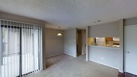 This single-floor residence offers an open, functional layout that connects the main living areas seamlessly. The living room is carpeted and bathed in natural light from a set of sliding glass doors dressed with vertical blinds, which lead to an outdoor area. The textured popcorn ceiling adds a classic touch throughout the space.

Adjacent to the living room, a defined dining area sits beneath a pendant light fixture, creating a clear sense of purpose within the open floor plan. The transition between these two areas is smooth, with consistent neutral-toned carpet and gray walls providing a cohesive, understated aesthetic.

The kitchen is positioned at the rear of the main living space and is accessible via a pass-through counter, allowing visual connectivity between cooking and living areas. The kitchen features under-cabinet lighting, light-toned cabinetry, and a refrigerator visible from the main space, offering a bright and practical cooking environment.

A hallway leads from the main living area toward additional rooms, including what appears to be a bathroom and at least one bedroom, completing this straightforward and efficiently designed single-floor layout.