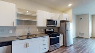 This single-floor unit presents a cohesive modern design with wood-style plank flooring that runs consistently throughout the space, creating a unified and polished aesthetic. The kitchen serves as a functional centerpiece, outfitted with crisp white cabinetry featuring bar-pull hardware, dark countertops, and a full suite of stainless steel appliances including a gas range, over-the-range microwave, and refrigerator. A tile backsplash adds texture behind the cooking area, while open shelving above the sink provides additional display or storage space. The bathroom mirrors the kitchen's contemporary styling with matching white cabinetry, dark countertops, an undermount sink, and a well-lit vanity mirror. The living area offers a generously sized, open layout with neutral gray walls, recessed lighting, and a ceiling fan in the bedroom area. A closet is accessible from the bedroom, and a window allows natural light to filter into the space while offering outside views. The overall floor plan, as seen from an aerial perspective, reflects an efficient and well-organized layout connecting the kitchen, living, sleeping, and bathroom areas in a compact yet practical arrangement.