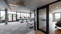 This fitness center is designed with a distinctly industrial-modern aesthetic, combining raw architectural elements with contemporary finishes to create a functional and visually dynamic workout environment. The space is organized into clearly defined zones, each tailored to a specific type of training.

The main gym floor features exposed concrete ceilings with visible ductwork painted dark, contributing to the industrial character of the space. Striped, multi-tonal carpet tiles in shades of gray, blue, and yellow cover the workout floor, providing both durability and visual interest. Large floor-to-ceiling windows and glass-paneled doors line one wall, flooding the cardio zone with natural light and offering views of the surrounding neighborhood. Treadmills, ellipticals, stationary bikes, and cable strength machines are thoughtfully arranged throughout the space, with full-length mirrors along one wall enhancing the sense of openness.

A separate, enclosed cycling studio provides a quieter training environment, featuring smooth hardwood-style flooring, recessed lighting, a clean white-and-gray palette, and a wall-mounted display screen. Matrix cycling bikes are arranged in rows, making the room well-suited for group or individual sessions.
