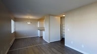 This single-floor space offers a clean, functional layout designed to maximize openness and ease of living. The main living and dining area flows seamlessly together, anchored by rich, dark hardwood-style flooring that extends throughout the unit. A stylish pendant light hangs over the dining area, adding a warm focal point to the space. Neutral gray walls and white trim create a bright, modern aesthetic that complements the overall design.

The kitchen is positioned off the main living area, separated by a partial wall that maintains an open feel while defining the cooking space. White cabinetry provides ample storage, and the layout is efficient and practical for everyday use. A ceiling-mounted light fixture keeps the kitchen well-lit.

Adjacent to the kitchen area, a built-in open shelving unit offers additional storage and display space, thoughtfully integrated into the architecture of the unit. This nook adds both character and utility to the layout.

The space is finished with clean white baseboards, neutral paint tones, and flush-mount lighting, giving it a well-maintained, move-in-ready appearance. The overall ambiance is calm and understated, making it a versatile space suited to a variety of living arrangements.