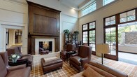 This expansive single-floor space is thoughtfully designed to accommodate a variety of social and recreational activities, combining classic architectural details with comfortable, functional furnishings throughout. The centerpiece of the main lounge is a dramatic two-story great room featuring a floor-to-ceiling wood-paneled fireplace with a stone surround, surrounded by rich brown leather seating arranged atop a plaid-patterned carpet. Soaring ceilings and tall French doors flood the area with natural light and provide direct access to an outdoor terrace. A grand piano positioned near the doors adds an elegant touch to this entertaining space.

Adjacent areas include a relaxed sitting room with cream upholstered sofas, recessed lighting, a wall-mounted television, and a second fireplace. A games area features a full-sized billiards table, built-in bookshelves, and a wall-mounted display screen. A dedicated dining room with dark hardwood floors showcases wainscoted walls and traditional wood dining tables. The fully equipped kitchen offers dark cabinetry, stone countertops, stainless steel appliances, and a seated island bar illuminated by pendant lighting. Decorative columns and wainscoting appear throughout, unifying the space with a consistently refined, traditional aesthetic.