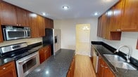 This single-story space centers around a galley-style kitchen that combines functionality with refined aesthetics. The kitchen is lined on both sides with warm, medium-toned wood cabinetry extending to the ceiling, offering generous storage throughout. Dark granite countertops with subtle speckled detailing run the length of both sides, providing ample workspace and a polished visual contrast against the cabinetry. A central island with matching granite continues the design language while enhancing prep space.

Stainless steel appliances, including a French door refrigerator with an in-door ice and water dispenser, a dishwasher, and an oven, lend a modern, cohesive look to the kitchen. Recessed lighting illuminates the space efficiently from above. Hardwood-style flooring runs through the kitchen, adding warmth underfoot.

A doorway at the far end of the kitchen leads into an adjoining room, where a classic chandelier is visible, hinting at a formal dining or living area. The neutral wall tones throughout create a clean, versatile backdrop, contributing to the overall bright and welcoming ambiance of the space.