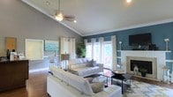 This single-floor home makes a strong first impression with its open-concept layout and thoughtfully curated design. The main living area is anchored by soaring vaulted ceilings that amplify the sense of spaciousness, while a striking teal accent wall adds bold contrast against the soft gray walls and crisp white trim. A ceiling fan with a light fixture keeps the space comfortable and well-lit, complemented by recessed lighting throughout. The living room is furnished with sleek white leather sofas arranged around a modern glass coffee table, resting atop a cowhide-style area rug over rich hardwood flooring. A classic fireplace with a clean, light-toned surround serves as a refined focal point on one end of the room, while a wall-mounted television anchors the teal wall on the opposite side. Large French doors and floor-to-ceiling curtain panels flood the space with natural light and provide a seamless connection to the outdoor patio, where glimpses of lush greenery are visible. A tall indoor plant adds a lively, organic touch to the interior. Just off the main living area, a casual dining or bar-height seating nook is visible, accented by colorful wall art, rounding out a home that balances modern aesthetics with warm, inviting character.