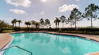 This single-story property is highlighted by a standout outdoor amenity: a large, sparkling swimming pool with clear turquoise water that immediately draws the eye. The pool area is framed by a well-maintained landscape of mature palm trees and tall pine trees, evoking a classic Florida setting. A paved pool deck surrounds the water, offering space for lounging, while neatly trimmed hedges and decorative plantings define the perimeter, providing a sense of privacy and enclosure. A fenced boundary at the rear opens to a broader tree-lined horizon, allowing for pleasant open views of the surrounding greenery beneath a bright, partly cloudy sky. The overall outdoor atmosphere is bright, airy, and inviting, characteristic of warm-weather living. The interior spaces of the property are part of the single-floor layout, rounding out a setting that balances comfortable indoor living with an appealing outdoor recreational area. The combination of the expansive pool, tropical landscaping, and open sky creates an ambiance well-suited for relaxation and outdoor enjoyment throughout the year.