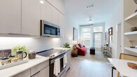 This contemporary single-floor residence offers a thoughtfully designed open-concept layout that seamlessly connects the kitchen and living areas. The kitchen is outfitted with sleek, flat-front cabinetry in a light wood and white finish, stainless steel appliances including a refrigerator, dishwasher, electric range, and over-the-range microwave, complemented by clean white quartz countertops and a classic subway tile backsplash. Light wood-tone flooring runs throughout, adding warmth and continuity across the space.

The living area is bright and airy, benefiting from soaring ceilings and floor-to-ceiling windows that flood the room with natural light. A glass-paned door opens directly onto a private balcony, offering outdoor views and additional ventilation. A ceiling fan adds both style and comfort to this welcoming gathering space.

The bedroom is a serene retreat, featuring a large window with sheer curtains that allow soft, diffused light to enter. The room is well-proportioned with clean white walls and the same warm hardwood-style flooring found throughout the residence.

The bathroom is finished to a high standard, with a wide vanity featuring ample storage, quartz countertops, and light wood cabinetry. A dedicated walk-in closet is accessible directly from the bathroom, providing generous storage. The overall aesthetic is clean, modern, and cohesive throughout.