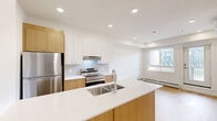 Located at 110 Lucas St NW in Calgary's growing community of Livingston, this thoughtfully designed one-bedroom unit offers a clean, contemporary living experience across a single floor. The open-concept layout maximizes the available space, creating a bright and airy atmosphere throughout.

The kitchen is a standout feature, finished with two-tone cabinetry combining warm wood-grain lower cabinets and crisp white upper cabinets. A large island with a white quartz countertop and an undermount sink serves as the central hub, complemented by stainless steel appliances including a refrigerator, electric range with oven, and an overhead range hood. Light wood-look flooring and recessed lighting complete the modern aesthetic.

The bedroom is a well-proportioned, sun-filled room with a window overlooking the neighbourhood streetscape. Baseboard heating, smooth white walls, and the same light hardwood-style flooring provide a clean and comfortable retreat.

The in-suite laundry area houses a stacked front-loading washer and dryer unit — a practical convenience rarely found in similarly sized homes. A private balcony extends the living space outdoors, offering views of the surrounding residential community. The overall design is bright, minimal, and move-in ready.