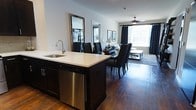 This single-floor residence offers a well-designed open-concept layout that seamlessly connects the kitchen, dining, and living areas into one cohesive living environment. The kitchen serves as the centerpiece of the space, anchored by a large island with a white countertop, an undermount sink with a modern brushed nickel faucet, dark cabinetry, and a stainless steel dishwasher. Rich, dark hardwood flooring runs throughout, adding warmth and continuity across all areas. The dining area is positioned just beyond the kitchen island, accommodating a full dining set with dark upholstered chairs, while a pair of tall framed mirrors along the wall enhance the sense of depth and openness. The living room benefits from generous natural light streaming through large windows dressed with dark curtains, creating a bright yet controllable atmosphere. A ceiling fan with a light fixture anchors the living area above, contributing to both comfort and style. The overall ambiance is modern and polished, with a cohesive dark-and-light contrast theme carried through the cabinetry, furnishings, and décor. The open flow between spaces makes this residence feel spacious, functional, and ideal for both everyday living and entertaining.