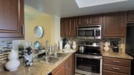 This single-story space showcases a thoughtfully designed kitchen that combines functionality with stylish finishes. Rich, dark wood cabinetry lines the walls, offering generous upper and lower storage with sleek bar-pull hardware. The granite countertops provide ample prep space and lend a polished, upscale feel to the room. A stainless steel double-basin sink is positioned conveniently along the counter, complemented by a modern pull-down faucet. The full suite of stainless steel appliances — including an electric range, over-the-range microwave, and additional countertop appliances — gives the kitchen a cohesive, contemporary aesthetic. A decorative mosaic tile backsplash in neutral tones with subtle color accents adds visual texture between the countertops and upper cabinetry. A skylight above introduces natural light, brightening the space and enhancing the warm tones of the wood and stone surfaces. Warm yellow-toned walls contribute to an inviting atmosphere throughout. The overall design balances practical kitchen functionality with refined decorative details, creating a space that is both efficient for everyday use and visually appealing.