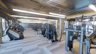 This modern fitness center is designed to accommodate a comprehensive range of workout routines within a single, expansive floor. The space is thoughtfully laid out to maximize usability, featuring rows of professional-grade strength training machines and cardio equipment arranged to allow easy movement between stations. The flooring consists of light-toned, neutral carpet tiles that provide both comfort underfoot and a clean, contemporary aesthetic throughout the space. Overhead, an exposed ceiling with industrial-style ductwork and suspended linear lighting fixtures creates a bright, evenly lit environment conducive to focused training. Structural columns are integrated seamlessly into the layout, framing distinct workout zones without disrupting the open flow of the space. Large mirrors are positioned along the walls, enhancing the sense of depth and allowing users to monitor their form during exercise. The overall ambiance is sleek and functional, with a dark equipment palette contrasting against the lighter tones of the flooring and walls. The facility reflects a high standard of fitness amenity, offering a well-organized, professional environment suited for both strength training and cardiovascular workouts.
