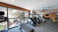 This fitness center is a thoughtfully designed workout facility that offers a comprehensive range of exercise options across a single, open floor plan. The space is immediately distinguished by its expansive floor-to-ceiling glass walls, which flood the interior with natural light and provide views of the surrounding outdoor area, creating an inviting and energizing atmosphere.

The cardio zone is prominently positioned along the windowed perimeter and features a lineup of treadmills, StairMaster steppers, and elliptical machines, all arranged to take advantage of the outdoor views. Structural columns clad in textured stone add an architectural accent to this section of the facility.

Moving deeper into the space, a dedicated strength training area houses a selection of resistance machines and cable equipment, complemented by a full rack of dumbbells in a range of weights. Large wall-to-wall mirrors amplify the sense of space and allow for proper form monitoring during workouts. A mounted flat-screen television and a ceiling fan enhance comfort in this zone.

Additional functional fitness elements include medicine balls mounted on a wall rack, a BOSU balance trainer, and open floor space suited for stretching or bodyweight exercises. Decorative wall art and warm terra-cotta accent walls contribute a polished, modern aesthetic throughout. Patterned carpet flooring, recessed lighting, and a glass-paneled entry door complete the well-appointed design of this fitness center.