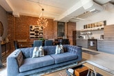 Located in Atlanta's vibrant 30308 corridor, this single-floor space masterfully blends industrial character with contemporary design. Exposed brick walls and polished concrete floors establish a raw, urban aesthetic that runs throughout, while curated furnishings and thoughtful finishes soften the industrial edge into something distinctly refined.

The living area anchors the space with large steel-framed windows that flood the room with natural light and frame views of the surrounding cityscape. A tufted sofa, industrial-style coffee table, and wall-mounted television are arranged against a deep charcoal accent wall, creating an inviting and stylish retreat.

The kitchen features sleek gray cabinetry, subway tile backsplash, stainless steel appliances including a gas range, open shelving, and a pass-through window that adds architectural interest and connectivity. Adjacent to the kitchen, a dedicated dining area with a solid wood table, leather chairs, and a statement chandelier is framed by exposed brick and bold geometric artwork.

The bedroom is a standout, featuring a striking yellow upholstered bed frame, an ornate chandelier, and exposed ductwork overhead that nods to the building's industrial roots. The bathroom continues the design narrative with brick walls, large-format tile flooring, a floating wood-tone vanity, and subway tile accents.