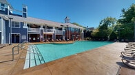 This community amenity facility offers a compelling dual-pool experience, combining a generous outdoor swimming pool with a fully enclosed indoor pool, each serving distinct purposes throughout the year. The outdoor pool is the centerpiece of the complex, featuring a wide, curvilinear design with a stamped concrete pool deck lined with lounge chairs for sunbathing. The adjoining clubhouse building is architecturally distinguished by its brick and dark blue siding exterior, a cupola-topped roofline, covered upper balconies with ceiling fans, and decorative columns — lending the structure a classic, resort-like character. Mature trees surround the outdoor area, providing natural shade and a lush backdrop under open skies. The indoor pool occupies a striking, purpose-built enclosure with soaring ceilings and an octagonal architectural form. Abundant natural light pours in through multi-tiered windows positioned high along the walls, creating a bright and airy atmosphere year-round. The warm wood-paneled ceiling adds a refined touch to the interior, while the pool surround offers ample deck space. Utility rooms and safety equipment are neatly integrated into the design. Decorative potted plants accent the indoor pool area, adding a welcoming, living element to the space. Together, these facilities provide a comprehensive aquatic amenity suited for all seasons.